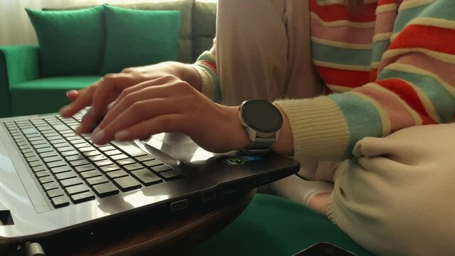a girl in a bright striped sweater sits on a green sofa and works with a laptop at home. typing on the keyboard close-up. watch on the girl's hand. there are many different things on the sofa. 