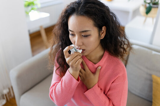 Asian Woman Using Inhaler While Suffering From Asthma At Home. Young Woman Using Asthma Inhaler. Close-up Of A Young Asian Woman Using Asthma Inhaler At Home.