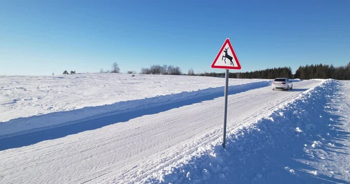 Road sign warning of the possible presence of wild animals on the way. A white car drives past a road sign on a winter sunny day. Country snowy road. Wildlife protection.Deer images on a road sign