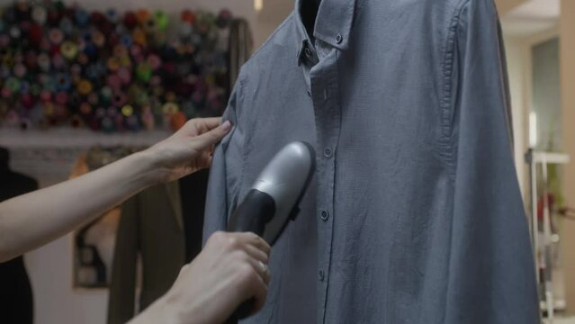 Close-up shot of woman irons tailored shirt with electric handheld steamer in atelier workshop. Male tailor sews clothes on background. Concept of small business, tailoring industry and fashion.
