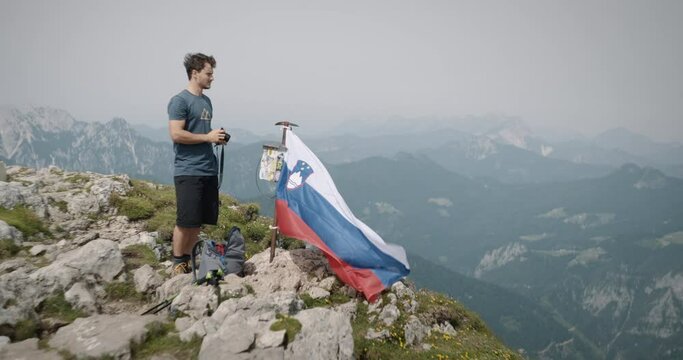 Hiker standing at the top of mountain Raduha, standing by the slovenina flag and his backpach tacking picture with a camera. Valley visible in background.
