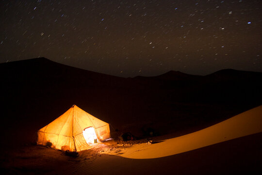 Star trails over a tent illuminated at night among sand dunes on a guided camel trek in Morocco.