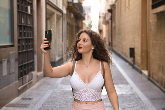 Young beautiful woman, blonde, with curly hair and blue eyes, wearing white top and pink skirt, on the street taking a selfie with her cell phone. Concept apps, smartphone, mobile.