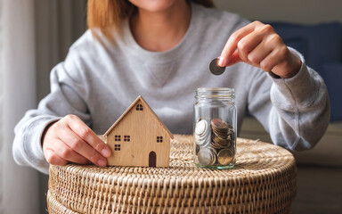 Closeup image of a woman putting coins into a glass jar with wooden house models for saving money...