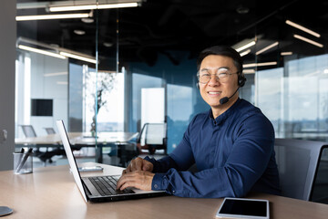 Portrait of successful office worker with headset for video call, asian man smiling and looking at camera working inside office, man in shirt and glasses customer service support.