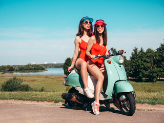 Fototapeta na wymiar Two young beautiful smiling hipster female in summer red bathing suits. Sexy carefree women driving retro motorbike. Positive models having fun, riding classic Italian scooter. In swimwear
