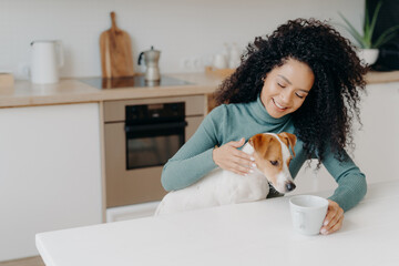Happy Afro African woman with curly hairstyle treats dog in kitchen, pose at white table with mug of drink, enjoy domestic atmosphere, have breakfast together. People, animals, home concept.