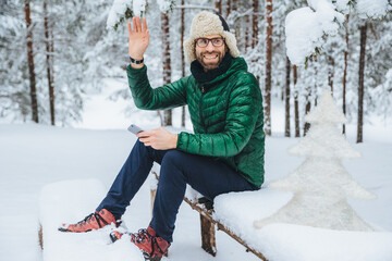 Outdoor shot of smiling male wears spectacles dressed in warm clothes, waves with hand as notices someone, holds smartphone, sits on wooden bench in winter forest, gestures actively
