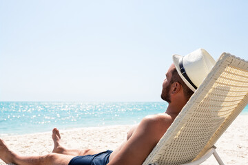 Young man in white hat relaxing in deck chair on beautiful sandy beach.