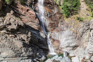 Bright view of flowing Lillaz waterfall (Cascate di Lillaz) with resulted pond beneath and granite karst rocks with evergreen trees, Aosta Valley, Italy