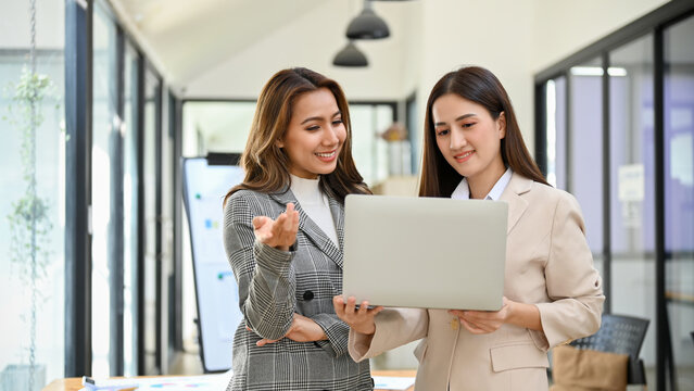 Two millennial Asian businesswomen looking at laptop screen while standing in the office.