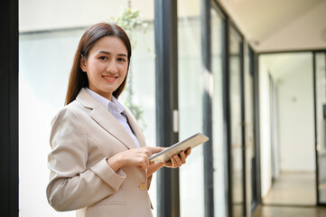 Charming Asian businesswoman stands in her office with tablet, smiling and looking at the camera.