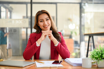 Gorgeous Asian business sits at her office desk, hands on chin, smiling and looking at the camera.