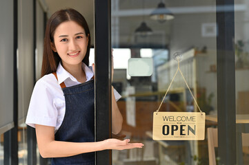 Beautiful young Asian female barista or coffee shop owner stands at her coffee shop's entrance door