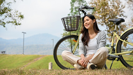 Relaxed Asian woman in casual clothes sits on the grass, taking a rest after ride a bike