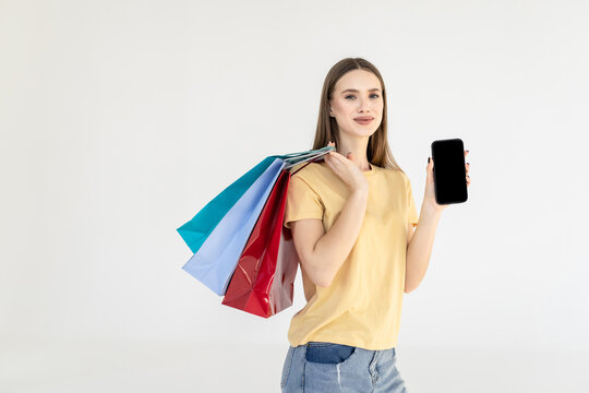 Happy woman standing with satisfied look showing shopping bags and smart phone with blank screen for advertisement isolated on white background.