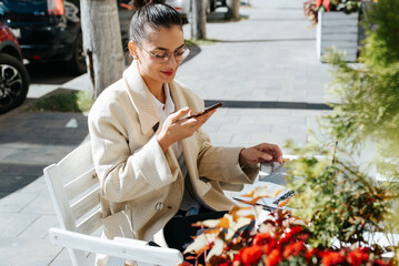 Smiling young brunette woman using voice assistant on smartphone while sitting at table with laptop working in street cafe on sunny day