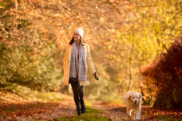 Woman Wearing Hat And Scarf With Pet Golden Retriever Dog On Walk Along Track In Autumn Countryside