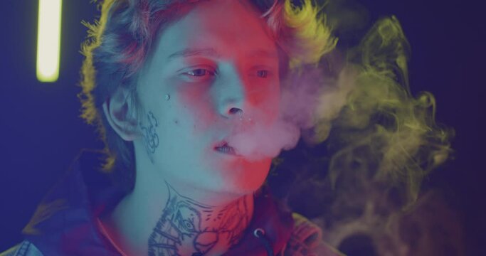 Close-up portrait of young man vaping exhaling smoke relaxing at night with neon lights in background. Addiction and youth lifestyle concept.