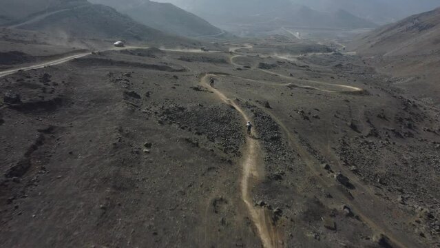 Drone shot of mountain bikers going downhill fast down a trail down hillside. A truck drives down a dirt path on left lifting up dirt in the air. Drone follows bikers while slowly decreasing altitude.