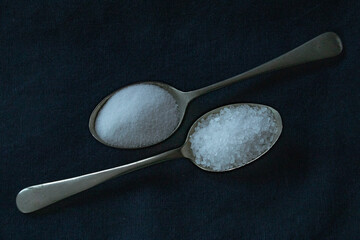 two silver spoons on a black background holding table salt and sea salt