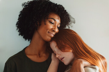 lesbian couple - African girl embracing young partner - red-haired Caucasian lady with closed-eyed, expressing love, affection, tenderness and a desire to be together - obrazy, fototapety, plakaty