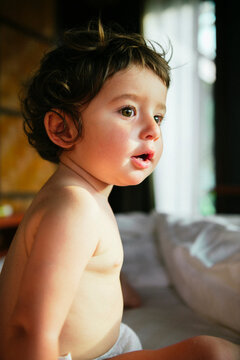 Side view of curious shirtless baby girl sitting on bed at home