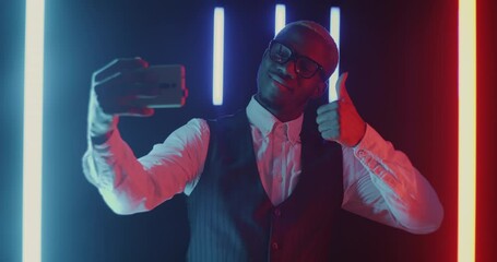 Slow motion of young businessman taking selfie with thumbs-up gesture using smartphone on black background with neon lamps. Modern lifestyle and successful businessconcept. - Powered by Adobe