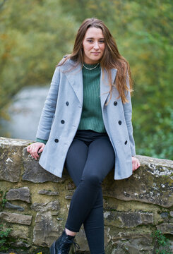 portrait of a young Caucasian woman with brown hair on a stone bridge with the green of the trees in the background. Dressed in a gray jacket, green sweater, dark pants and militar