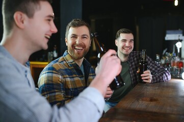 Three young men in casual clothes are smiling, holding bottles of beer while standing near bar counter in pub