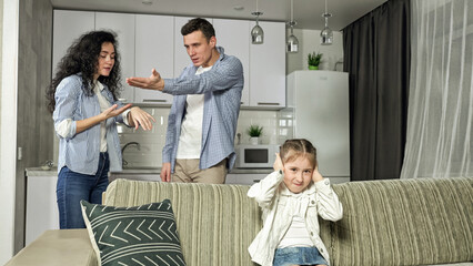 Fototapeta premium Junior schoolgirl sits closing ears with hands against parents arguing behind sofa. Angry father and mother quarrel and argue about daughter naughty behavior, closeup