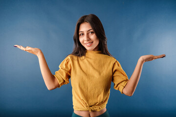 Portrait of cute dark-haired woman wearing casual top isolated over blue background shrugging and throwing hands aside with clueless. Not sure what to do. Looking at the camera.
