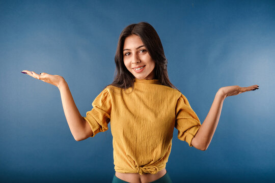 Portrait of cute dark-haired woman wearing casual top isolated over blue background shrugging and throwing hands aside with clueless. Not sure what to do. Looking at the camera.