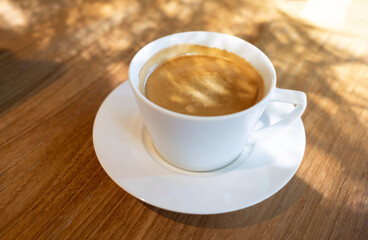 White cup of hot coffee on wooden desk with sunlight in the background in the morning time