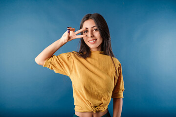 Cute dark-haired woman wearing casual top isolated over blue background doing peace symbol with fingers over face, smiling cheerful showing victory.