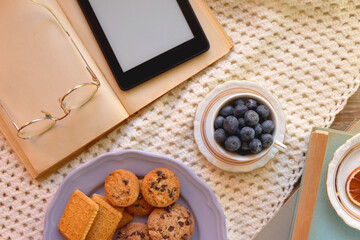 Plate of cookies, cup of tea, fresh blueberries, dry oranges, stack of books, reading glasses and tablet on the table. Hygge at home. Flat lay.