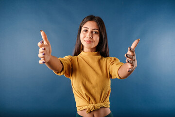 Beautiful brunette woman wearing mustard yellow t-shirt isolated over blue background looking at the camera smiling with open arms for hug. Cheerful expression embracing happiness. Got some good news.
