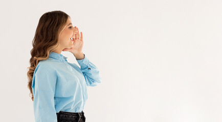 Hey! Cheerful young lady in blue shirt is shouting and holding hand near her open mouth on light...