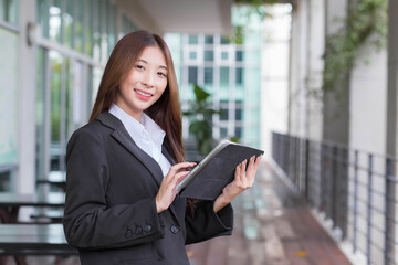 Asian professional business young woman in dark blue suit smiles happily stand with confidence and look at the camera while she works and holds tablet front office.