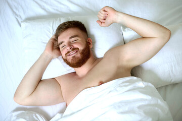 Happy young handsome man is waking up in the morning in bed in bedroom, stretching his arms, body, smiling.