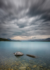 Long exposure image of two rocks in the turquoise blue water of Lake Tekapo in New Zealand with snow capped mountains in the distance and dramatic clouds overhead