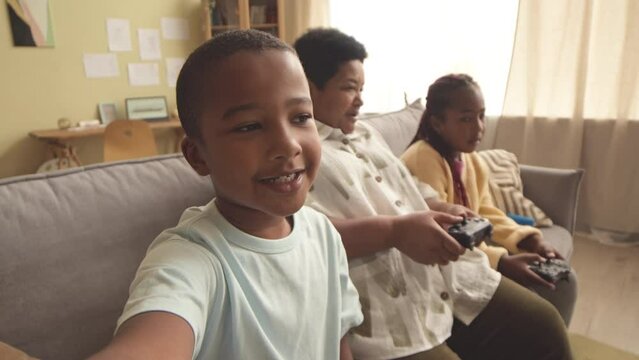 Cool African American tween boy recording video of himself spending leisure time at home with grandma and elder sister playing video games sitting on sofa in living room using controller