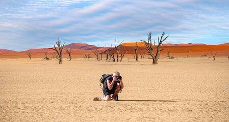 Fototapeta na wymiar Photographer sitting between dead trees in deadvlei during sunrise take a photo, famous natural landmark with full moon - Namib desert, Namibia 
