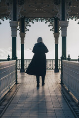 Brighton, The Bandstand, blue sky and clouds in winter with an adult female person silhouette, symmetrical archway, turned away, wearing hat and long coat