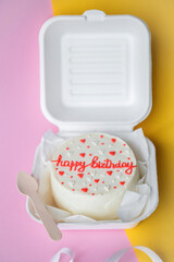 A small bento birthday cake in a box with a wooden spoon. Yellow and pink background