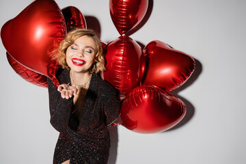 happy and blonde woman in black dress holding red heart-shaped balloons and gesturing on grey.