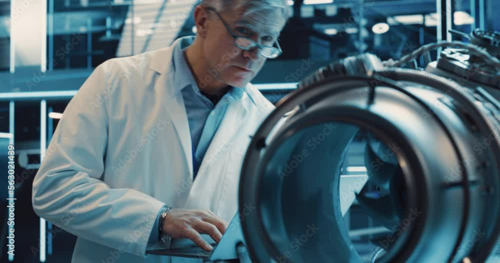 Poster middle aged industrial engineer working on a futuristic jet engine, standing with laptop computer in