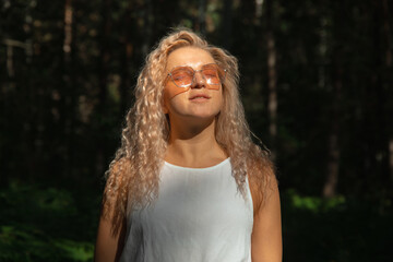 The young woman in sunglasses looking at the sun against the background of a dark forest