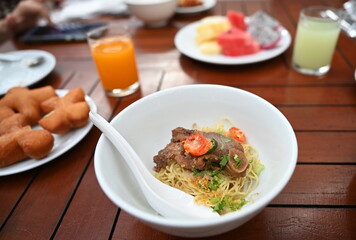 Noodles with braised pork without soup in white cup, plastic spoon. Yellow egg noodles with large chunks of stewed pork topped with sliced ​​coriander and seasoned with sugar, vinegar and chilli.  
