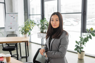Asian interior designer holding eyeglasses and looking at camera in office.
